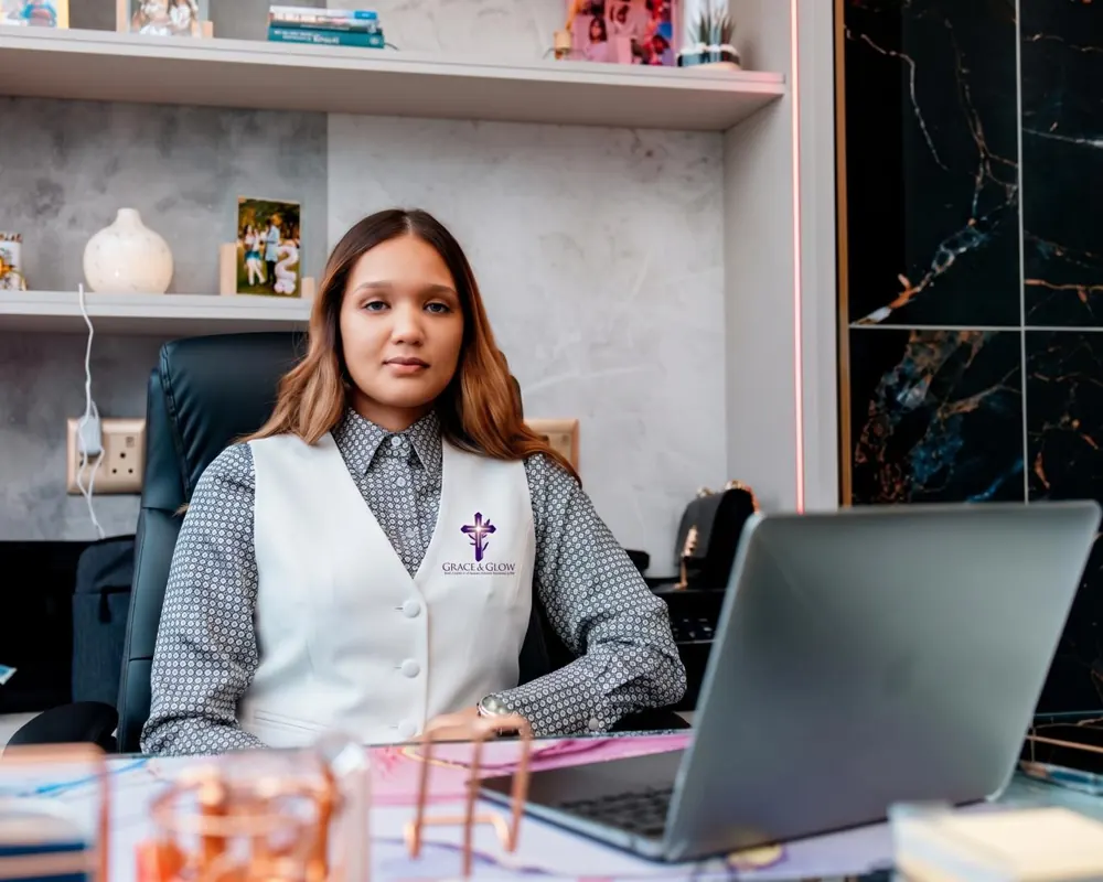 Kayla Fisher, founder of Grace & Glow, seated at her desk in a white vest bearing the Grace & Glow logo